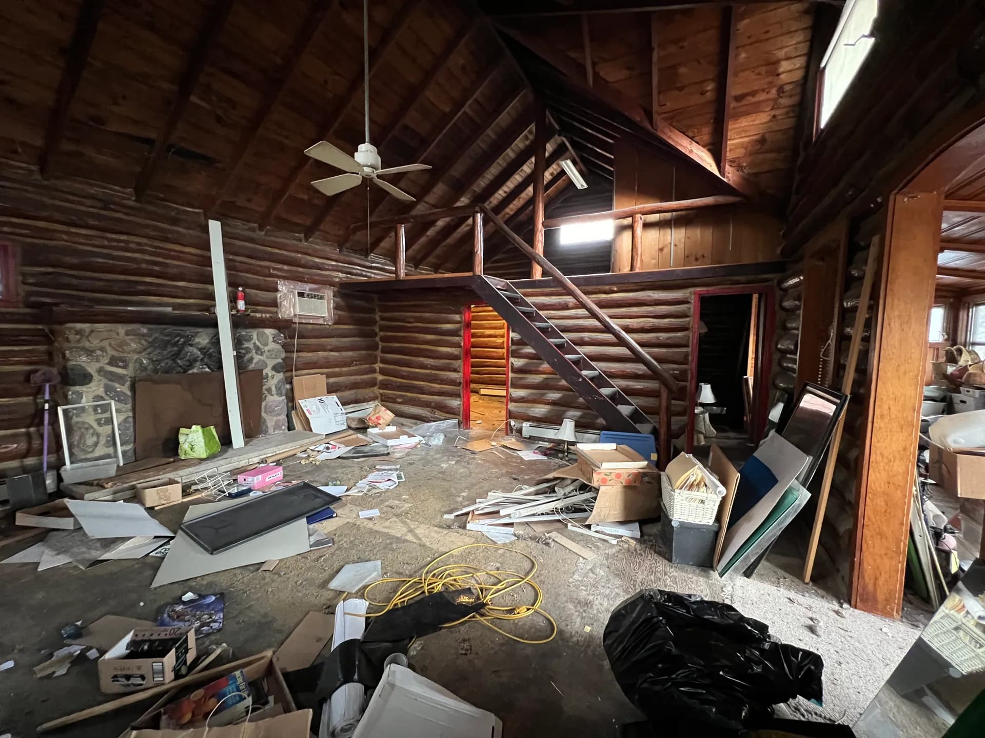 Debris-filled cabin living room before renovation — stone fireplace, loft, and log staircase